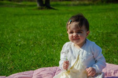 Parkta yeşil çim üzerinde pembe ekose parlak giysiler bebek. Yeni doğan bebek sıcak bir günde dışarıda. Küçük kız ilkbaharda yeşil bir çim enlaid üzerinde bir ekose üzerinde oynuyor