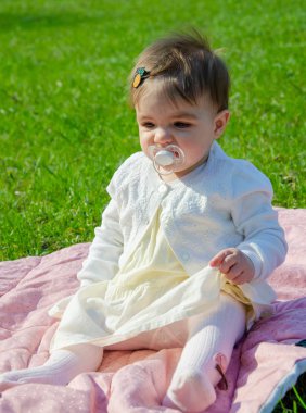 Parkta yeşil çim üzerinde pembe ekose parlak giysiler bebek. Yeni doğan bebek sıcak bir günde dışarıda. Küçük kız ilkbaharda yeşil bir çim enlaid üzerinde bir ekose üzerinde oynuyor
