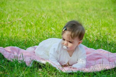 Parkta yeşil çim üzerinde pembe ekose parlak giysiler bebek. Yeni doğan bebek sıcak bir günde dışarıda. Küçük kız ilkbaharda yeşil bir çim enlaid üzerinde bir ekose üzerinde oynuyor