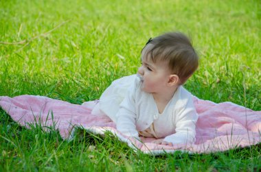 Parkta yeşil çim üzerinde pembe ekose parlak giysiler bebek. Yeni doğan bebek sıcak bir günde dışarıda. Küçük kız ilkbaharda yeşil bir çim enlaid üzerinde bir ekose üzerinde oynuyor