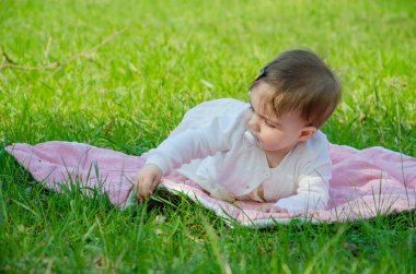 Parkta yeşil çim üzerinde pembe ekose parlak giysiler bebek. Yeni doğan bebek sıcak bir günde dışarıda. Küçük kız ilkbaharda yeşil bir çim enlaid üzerinde bir ekose üzerinde oynuyor