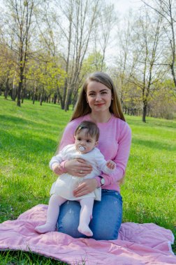 Yeşil sağ pembe ekose parlak giysiler içinde bebek ile anne. Sıcak bir günde parkta dinlenen aile. Anne ve küçük kız parkta 10 ay yürümek