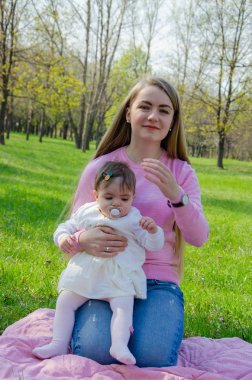 Yeşil sağ pembe ekose parlak giysiler içinde bebek ile anne. Sıcak bir günde parkta dinlenen aile. Anne ve küçük kız parkta 10 ay yürümek