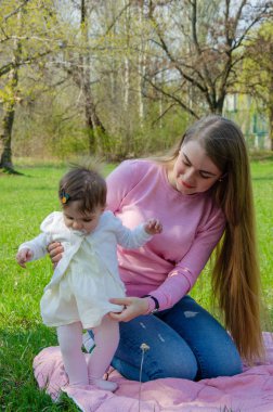 Yeşil sağ pembe ekose parlak giysiler içinde bebek ile anne. Sıcak bir günde parkta dinlenen aile. Anne ve küçük kız parkta 10 ay yürümek