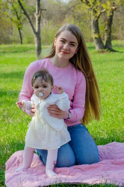 Yeşil sağ pembe ekose parlak giysiler içinde bebek ile anne. Sıcak bir günde parkta dinlenen aile. Anne ve küçük kız parkta 10 ay yürümek