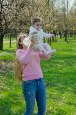 Yeşil sağ pembe ekose parlak giysiler içinde bebek ile anne. Sıcak bir günde parkta dinlenen aile. Anne ve küçük kız parkta 10 ay yürümek