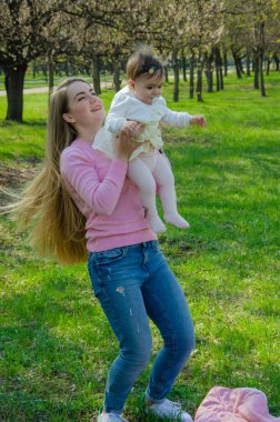 Yeşil sağ pembe ekose parlak giysiler içinde bebek ile anne. Sıcak bir günde parkta dinlenen aile. Anne ve küçük kız parkta 10 ay yürümek
