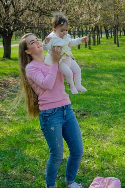 Yeşil sağ pembe ekose parlak giysiler içinde bebek ile anne. Sıcak bir günde parkta dinlenen aile. Anne ve küçük kız parkta 10 ay yürümek