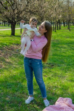 Yeşil sağ pembe ekose parlak giysiler içinde bebek ile anne. Sıcak bir günde parkta dinlenen aile. Anne ve küçük kız parkta 10 ay yürümek