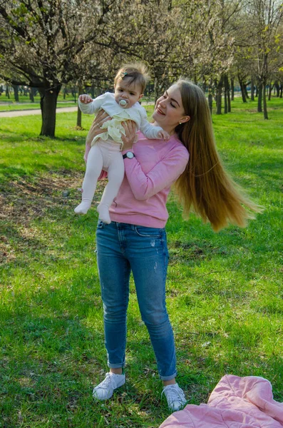 Yeşil sağ pembe ekose parlak giysiler içinde bebek ile anne. Sıcak bir günde parkta dinlenen aile. Anne ve küçük kız parkta 10 ay yürümek