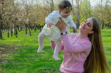 Yeşil sağ pembe ekose parlak giysiler içinde bebek ile anne. Sıcak bir günde parkta dinlenen aile. Anne ve küçük kız parkta 10 ay yürümek