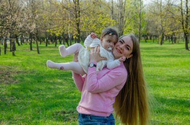 Yeşil sağ pembe ekose parlak giysiler içinde bebek ile anne. Sıcak bir günde parkta dinlenen aile. Anne ve küçük kız parkta 10 ay yürümek