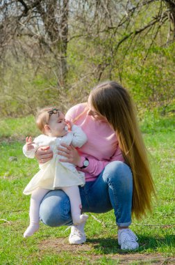 Yeşil sağ pembe ekose parlak giysiler içinde bebek ile anne. Sıcak bir günde parkta dinlenen aile. Anne ve küçük kız parkta 10 ay yürümek
