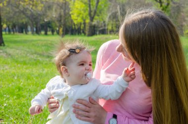 Yeşil sağ pembe ekose parlak giysiler içinde bebek ile anne. Sıcak bir günde parkta dinlenen aile. Anne ve küçük kız parkta 10 ay yürümek