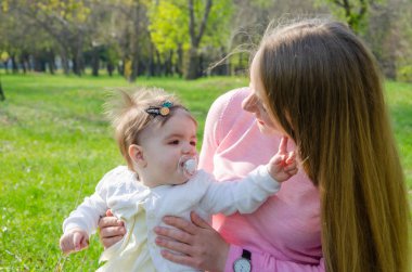 Yeşil sağ pembe ekose parlak giysiler içinde bebek ile anne. Sıcak bir günde parkta dinlenen aile. Anne ve küçük kız parkta 10 ay yürümek