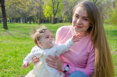 Yeşil sağ pembe ekose parlak giysiler içinde bebek ile anne. Sıcak bir günde parkta dinlenen aile. Anne ve küçük kız parkta 10 ay yürümek