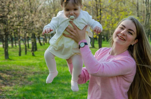 Mom with baby in bright clothes on a pink plaid on the green right. Family resting in the park on a warm day. Mom and little girl 10 months walk in the park