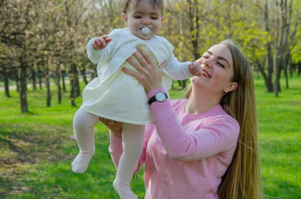 Mom with baby in bright clothes on a pink plaid on the green right. Family resting in the park on a warm day. Mom and little girl 10 months walk in the park