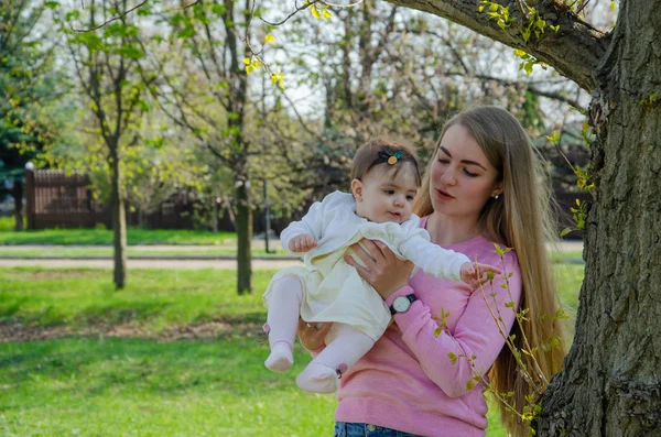 Mom with baby in bright clothes on a pink plaid on the green right. Family resting in the park on a warm day. Mom and little girl 10 months walk in the park