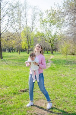 Yeşil sağ pembe ekose parlak giysiler içinde bebek ile anne. Sıcak bir günde parkta dinlenen aile. Anne ve küçük kız parkta 10 ay yürümek