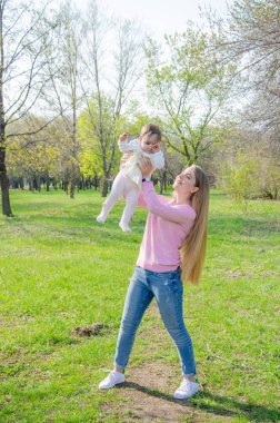 Yeşil sağ pembe ekose parlak giysiler içinde bebek ile anne. Sıcak bir günde parkta dinlenen aile. Anne ve küçük kız parkta 10 ay yürümek
