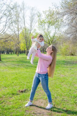 Yeşil sağ pembe ekose parlak giysiler içinde bebek ile anne. Sıcak bir günde parkta dinlenen aile. Anne ve küçük kız parkta 10 ay yürümek