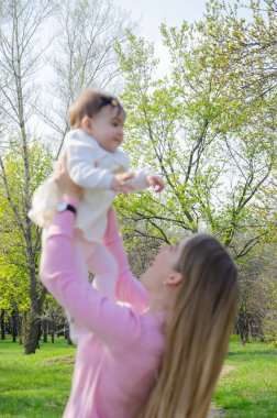Yeşil sağ pembe ekose parlak giysiler içinde bebek ile anne. Sıcak bir günde parkta dinlenen aile. Anne ve küçük kız parkta 10 ay yürümek