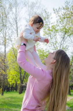 Yeşil sağ pembe ekose parlak giysiler içinde bebek ile anne. Sıcak bir günde parkta dinlenen aile. Anne ve küçük kız parkta 10 ay yürümek