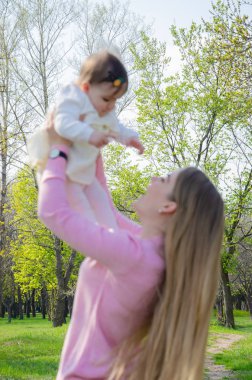 Yeşil sağ pembe ekose parlak giysiler içinde bebek ile anne. Sıcak bir günde parkta dinlenen aile. Anne ve küçük kız parkta 10 ay yürümek