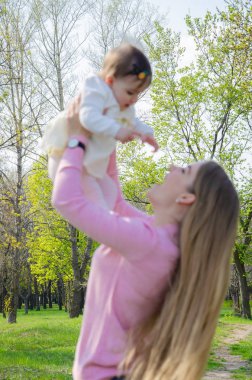 Yeşil sağ pembe ekose parlak giysiler içinde bebek ile anne. Sıcak bir günde parkta dinlenen aile. Anne ve küçük kız parkta 10 ay yürümek