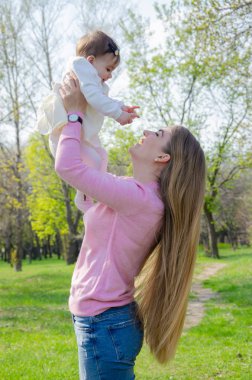 Yeşil sağ pembe ekose parlak giysiler içinde bebek ile anne. Sıcak bir günde parkta dinlenen aile. Anne ve küçük kız parkta 10 ay yürümek