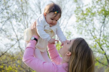 Yeşil sağ pembe ekose parlak giysiler içinde bebek ile anne. Sıcak bir günde parkta dinlenen aile. Anne ve küçük kız parkta 10 ay yürümek