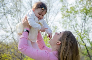 Yeşil sağ pembe ekose parlak giysiler içinde bebek ile anne. Sıcak bir günde parkta dinlenen aile. Anne ve küçük kız parkta 10 ay yürümek