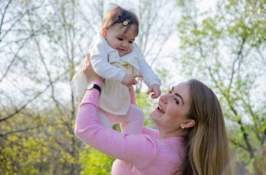Yeşil sağ pembe ekose parlak giysiler içinde bebek ile anne. Sıcak bir günde parkta dinlenen aile. Anne ve küçük kız parkta 10 ay yürümek