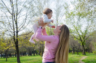 Yeşil sağ pembe ekose parlak giysiler içinde bebek ile anne. Sıcak bir günde parkta dinlenen aile. Anne ve küçük kız parkta 10 ay yürümek