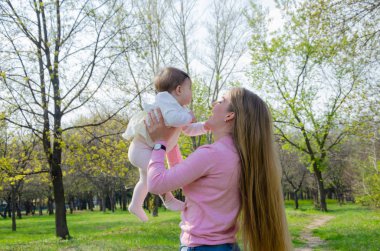 Yeşil sağ pembe ekose parlak giysiler içinde bebek ile anne. Sıcak bir günde parkta dinlenen aile. Anne ve küçük kız parkta 10 ay yürümek
