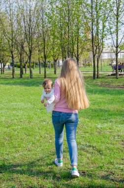 Yeşil sağ pembe ekose parlak giysiler içinde bebek ile anne. Sıcak bir günde parkta dinlenen aile. Anne ve küçük kız parkta 10 ay yürümek