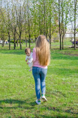 Yeşil sağ pembe ekose parlak giysiler içinde bebek ile anne. Sıcak bir günde parkta dinlenen aile. Anne ve küçük kız parkta 10 ay yürümek