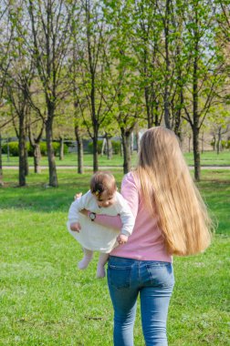 Yeşil sağ pembe ekose parlak giysiler içinde bebek ile anne. Sıcak bir günde parkta dinlenen aile. Anne ve küçük kız parkta 10 ay yürümek