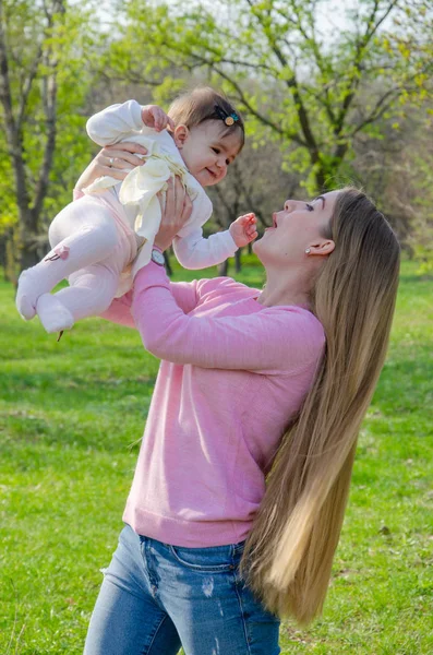 Mom with baby in bright clothes on a pink plaid on the green right. Family resting in the park on a warm day. Mom and little girl 10 months walk in the park