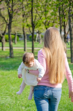 Yeşil sağ pembe ekose parlak giysiler içinde bebek ile anne. Sıcak bir günde parkta dinlenen aile. Anne ve küçük kız parkta 10 ay yürümek