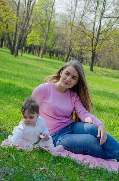 Mom with baby in bright clothes on a pink plaid on the green right. Family resting in the park on a warm day. Mom and little girl 10 months walk in the park