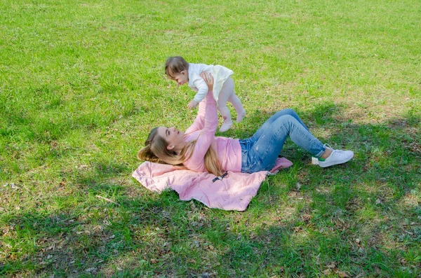 Mom with baby in bright clothes on a pink plaid on the green right. Family resting in the park on a warm day. Mom and little girl 10 months walk in the park