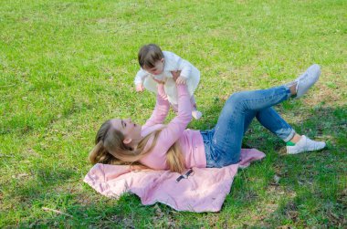 Yeşil sağ pembe ekose parlak giysiler içinde bebek ile anne. Sıcak bir günde parkta dinlenen aile. Anne ve küçük kız parkta 10 ay yürümek