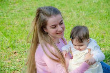 Yeşil sağ pembe ekose parlak giysiler içinde bebek ile anne. Sıcak bir günde parkta dinlenen aile. Anne ve küçük kız parkta 10 ay yürümek