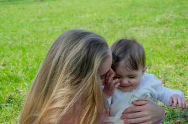 Yeşil sağ pembe ekose parlak giysiler içinde bebek ile anne. Sıcak bir günde parkta dinlenen aile. Anne ve küçük kız parkta 10 ay yürümek