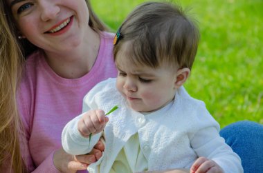 Yeşil sağ pembe ekose parlak giysiler içinde bebek ile anne. Sıcak bir günde parkta dinlenen aile. Anne ve küçük kız parkta 10 ay yürümek