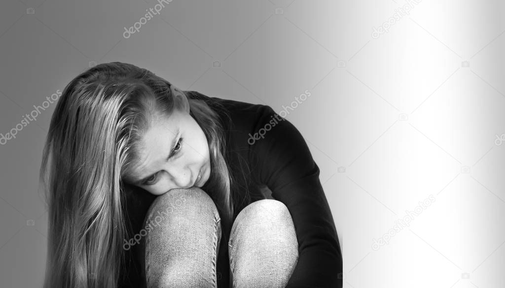 Sad beautiful girl with long blonde hair dressed in jeans and a black sweater rested her head on her bent knees, photo in black and white format on a white background