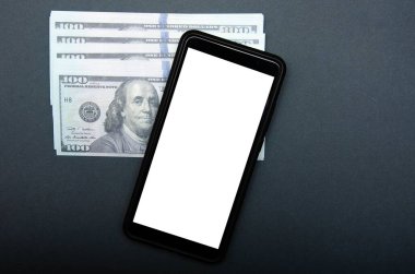 Dolar ve akıllı telefon. Üzerlerinde 100 dolarlık banknotlar var. Yanlarında beyaz ekranlı bir akıllı telefon var. Üst manzara ve açı. Makro akıllı telefon ve dolar. 