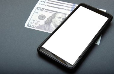 Dolar ve akıllı telefon. Üzerlerinde 100 dolarlık banknotlar var. Yanlarında beyaz ekranlı bir akıllı telefon var. Üst manzara ve açı. Makro akıllı telefon ve dolar. 